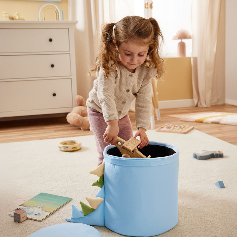 Otrok pospravlja lesene igrače v modro škatlo za shranjevanje z motivom dinozavra v spalnici.