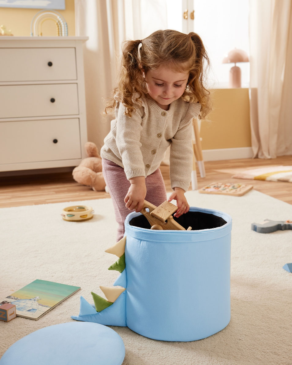 Otrok pospravlja lesene igrače v modro škatlo za shranjevanje z motivom dinozavra.