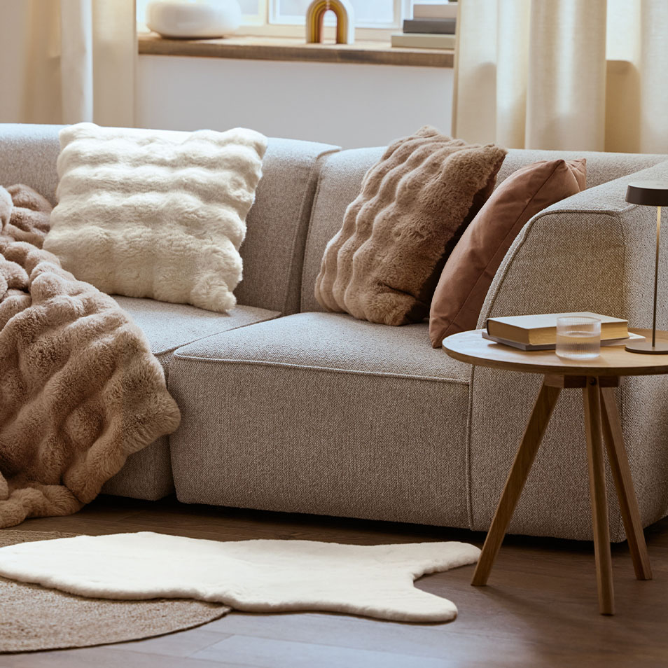 Siva modularna zofa z mehkimi blazinami in odejo, poleg lesene stranske mizice.