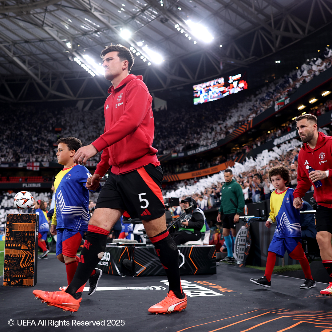 Nogometaši v rdečih dresih in črnih kratkih hlačah hodijo po igrišču z otroki, oblečenimi v športna oblačila.