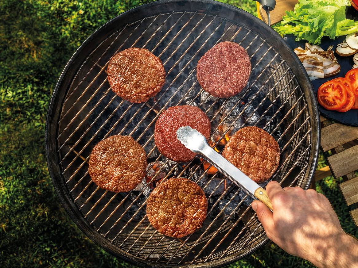 Peka burgerjev na žaru na prostem, s svežo zelenjavo ob strani.