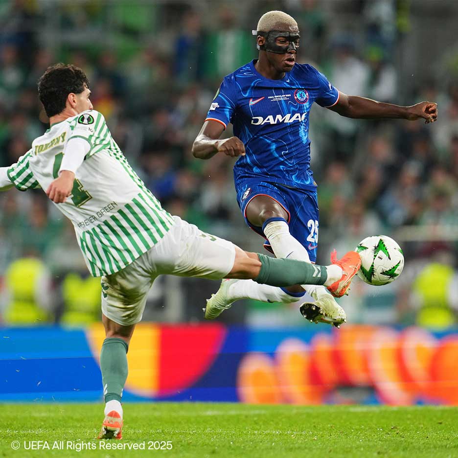 Dva nogometaša se borita za žogo na igrišču, eden v belo-zelenem, drugi v modrem z masko.