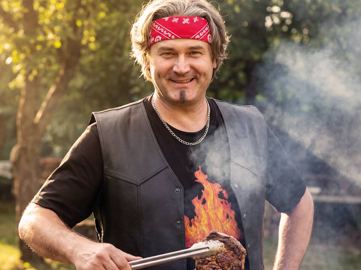 Moški v usnjenem brezrokavniku in rdeči ruti peče zrezek na žaru.