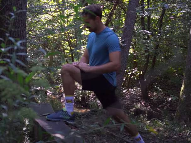 Moški v modri majici in črnih kratkih hlačah se razteza v gozdu.