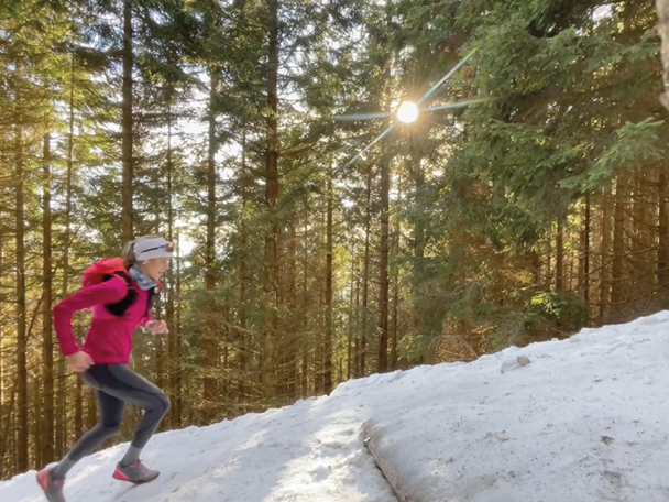 Ženska teče po zasneženi gozdni poti v rožnati jakni in s čelno svetilko.