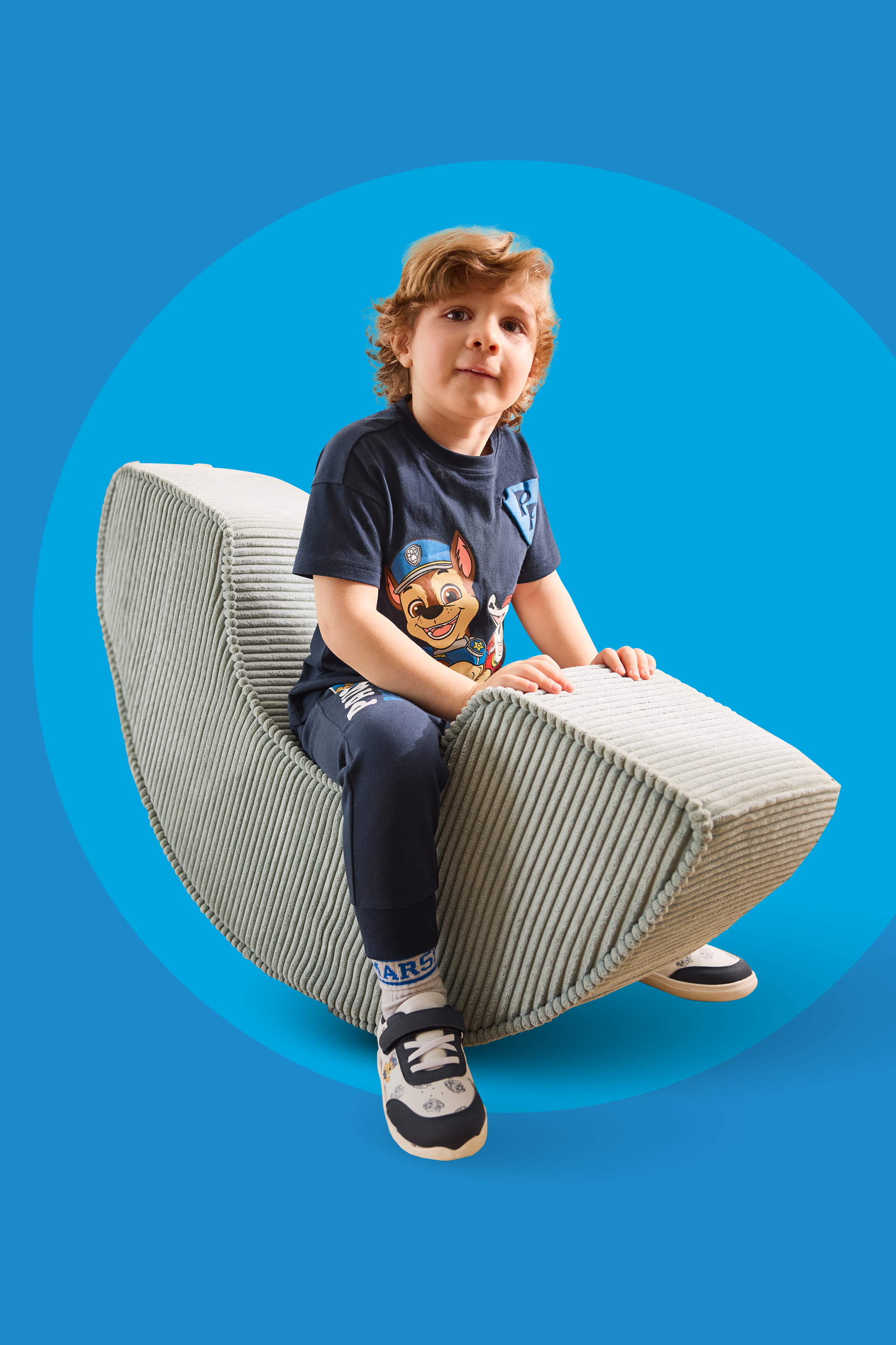 Deček v modri majici s potiskom in hlačah sedi na sivi blazini.