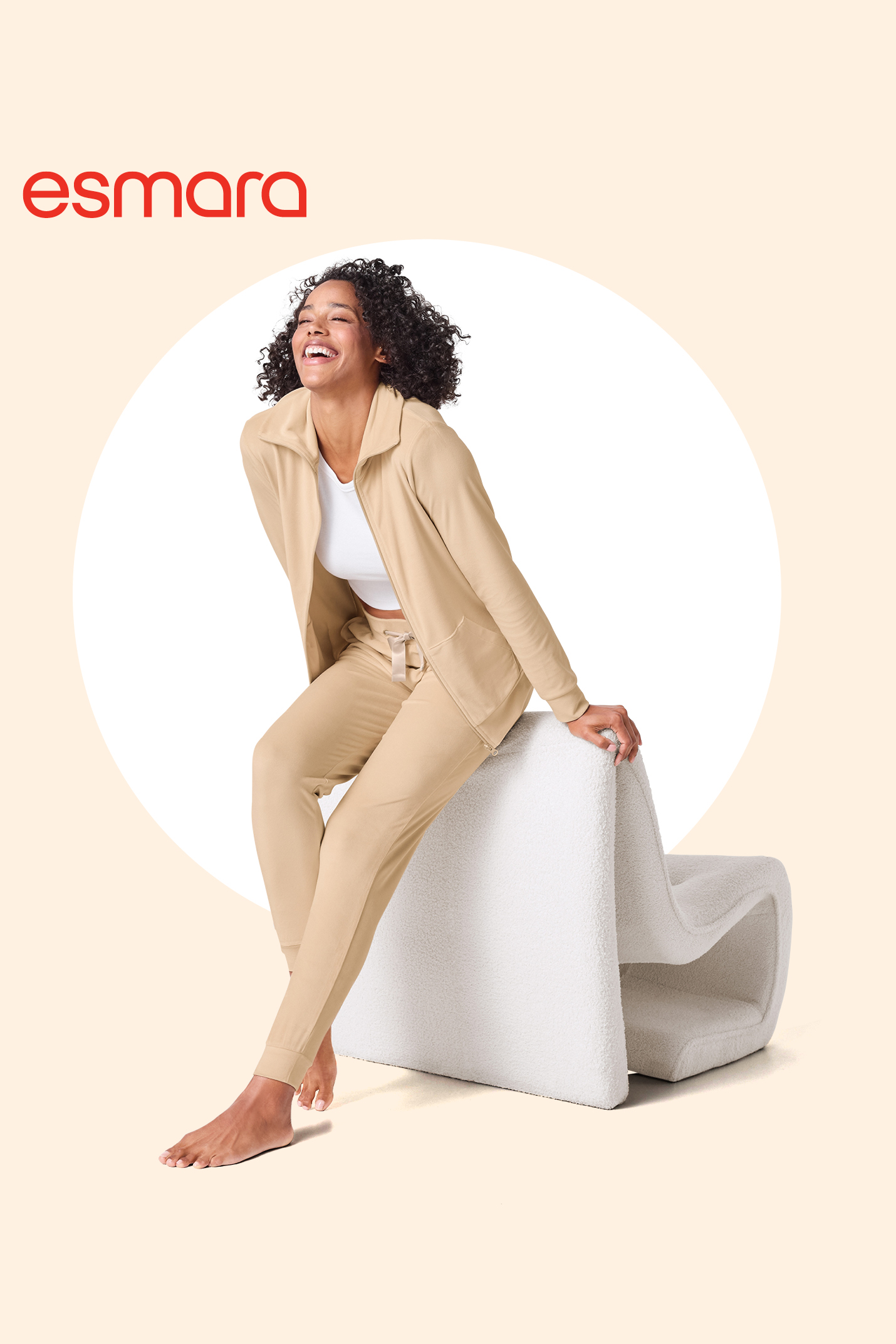 Ženska v bež trenirki in beli majici, smejoča se, sedi na belem stolu.