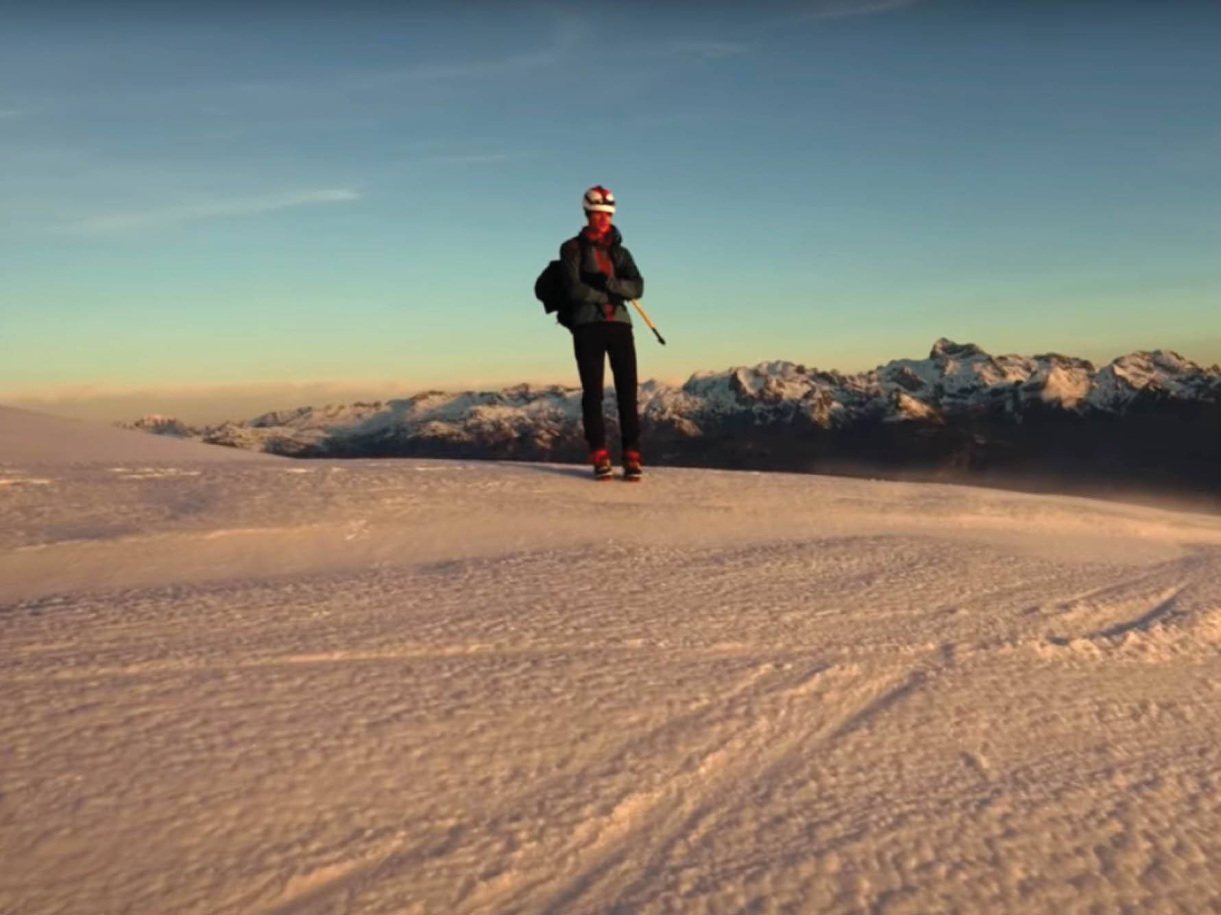 Planinec stoji na zasneženem vrhu gore z razgledom na zasnežene gore v ozadju.