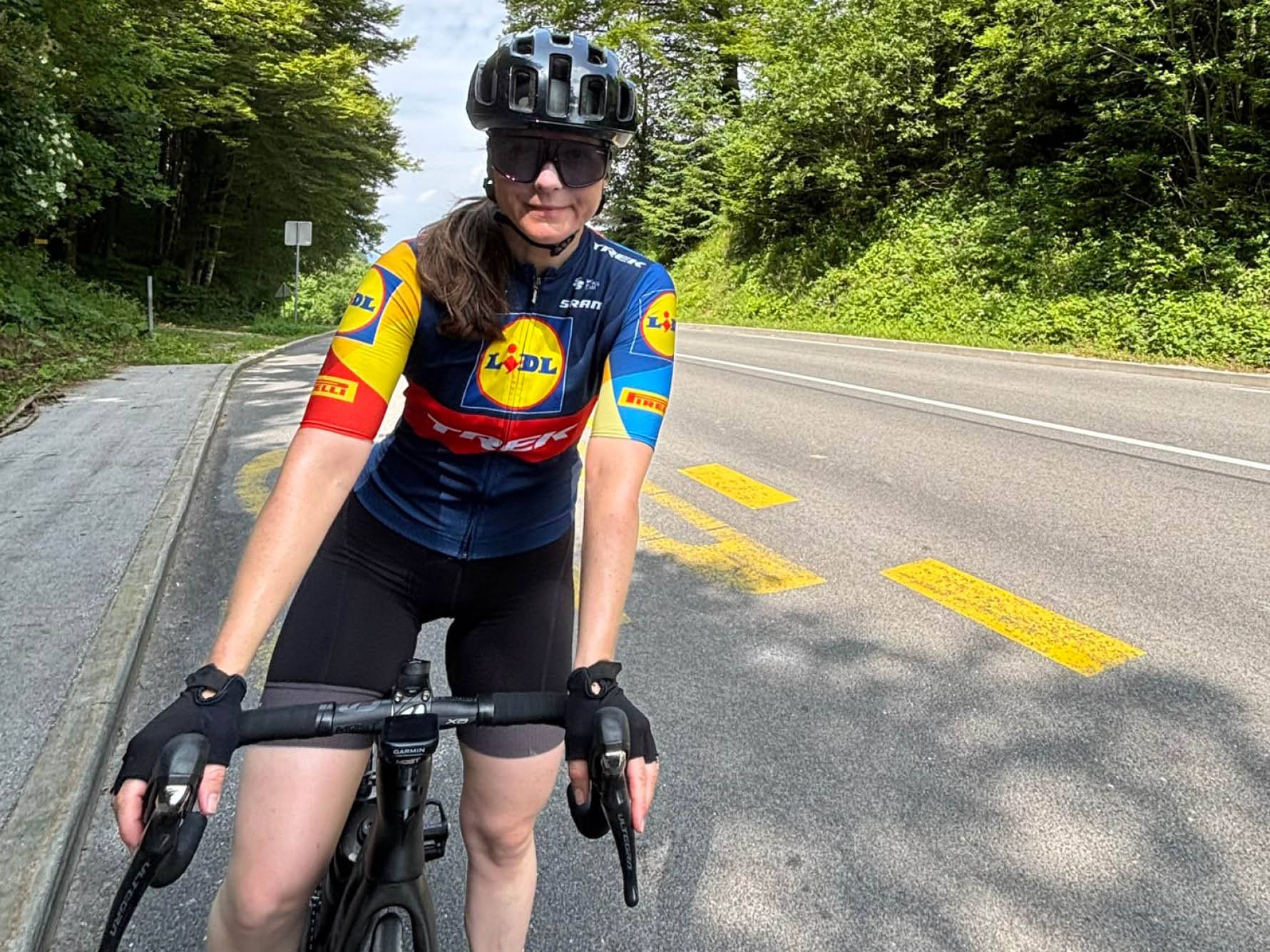 Kolesarka v dresu Lidl Trek kolesari po cesti, obdana z zelenjem.