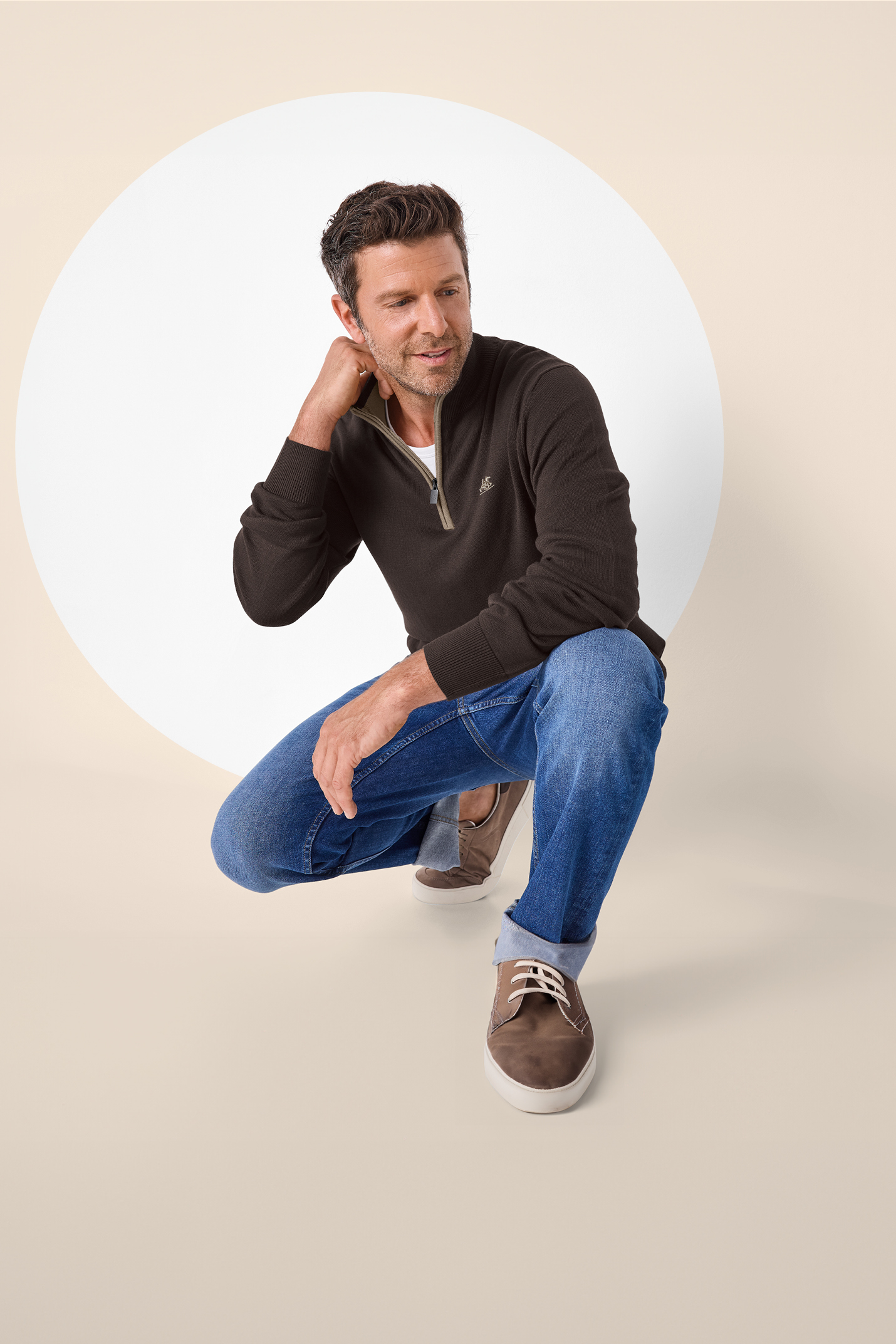 Moški v temno rjavem puloverju z zadrgo, modrih kavbojkah in rjavih čevljih.