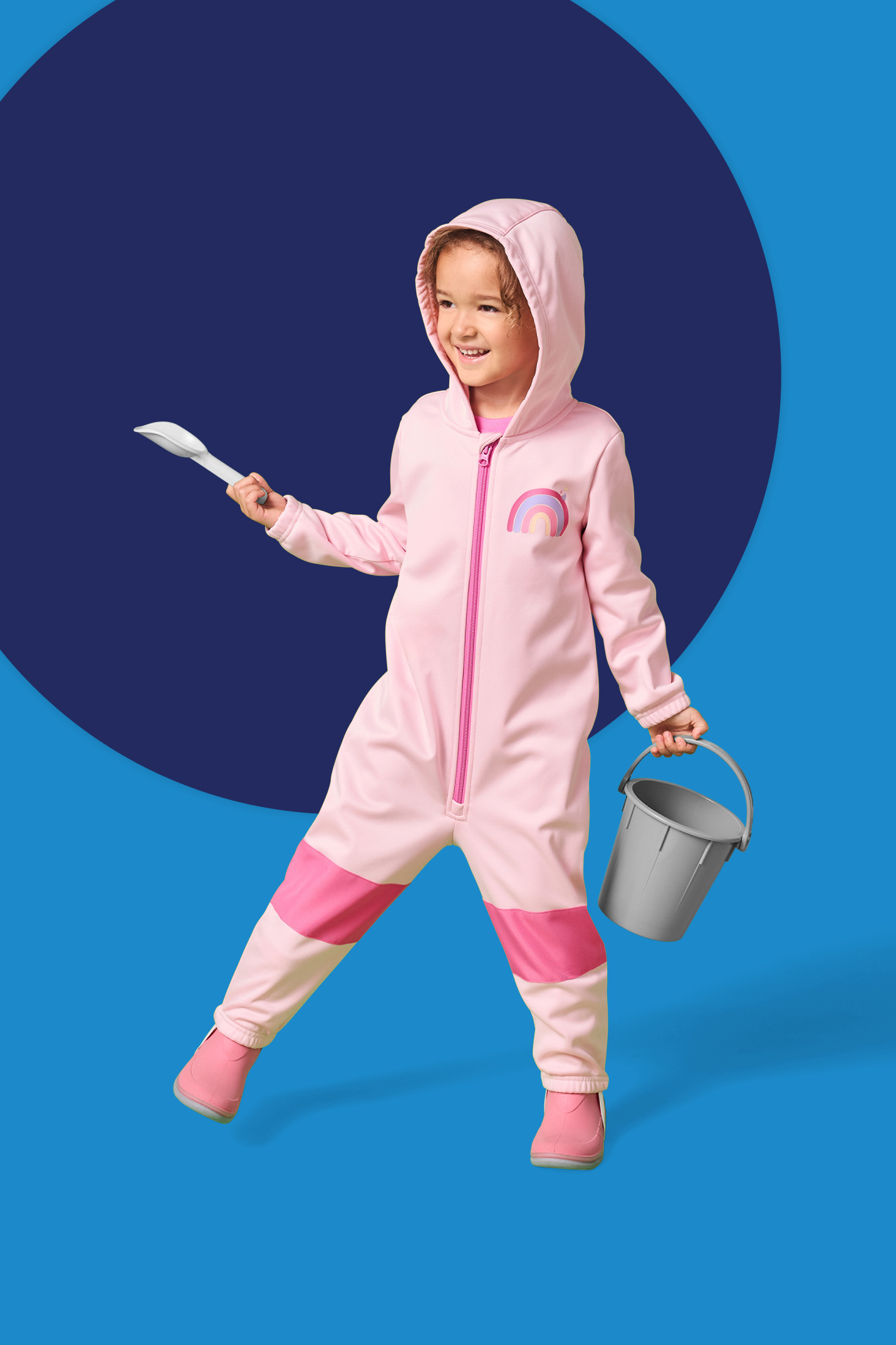 Deklica v rožnatem kombinezonu z mavrico, drži lopatko in vedro.