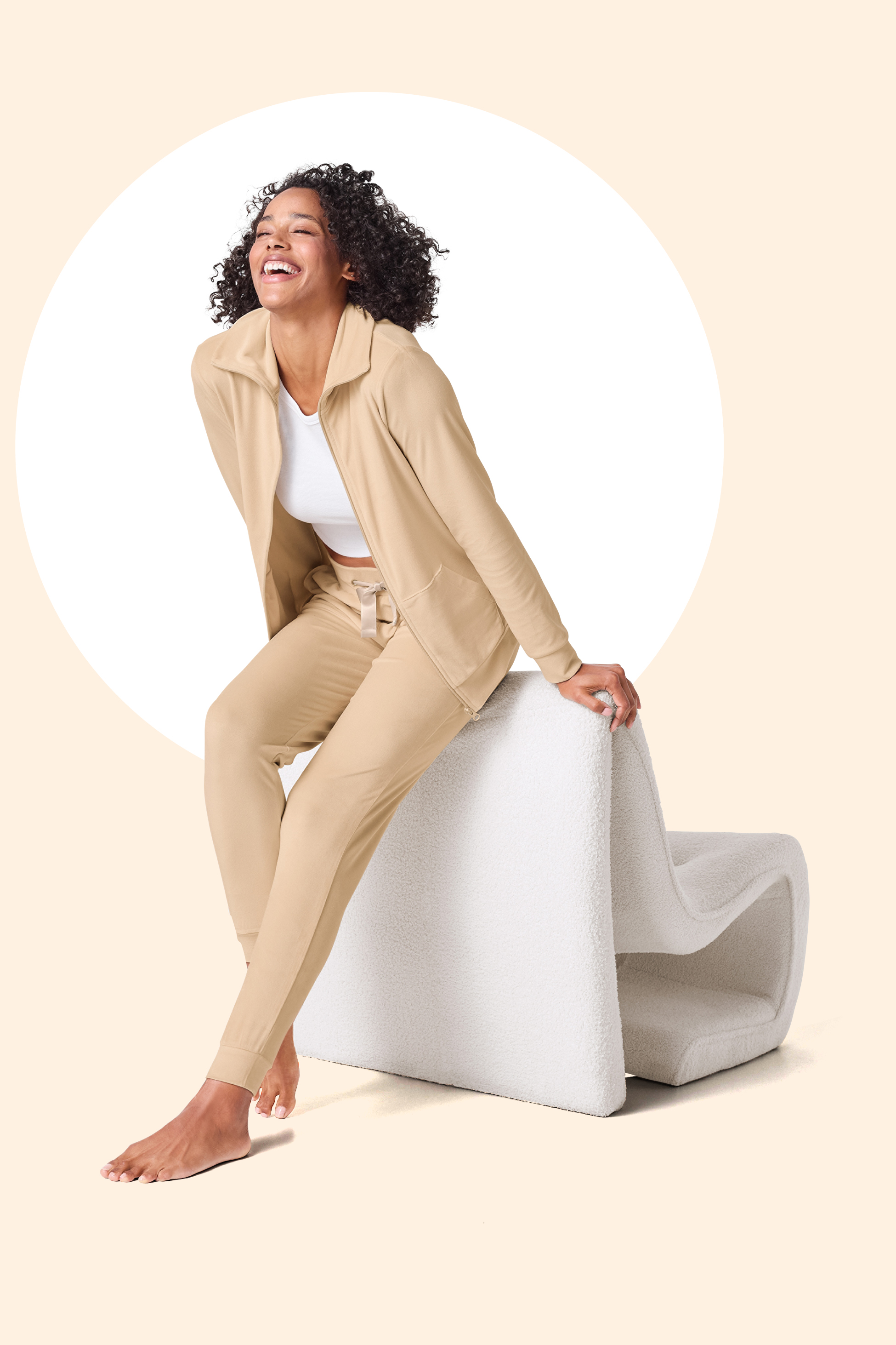 Ženska v bež trenirki in beli majici se smehlja, sedi na belem stolu.